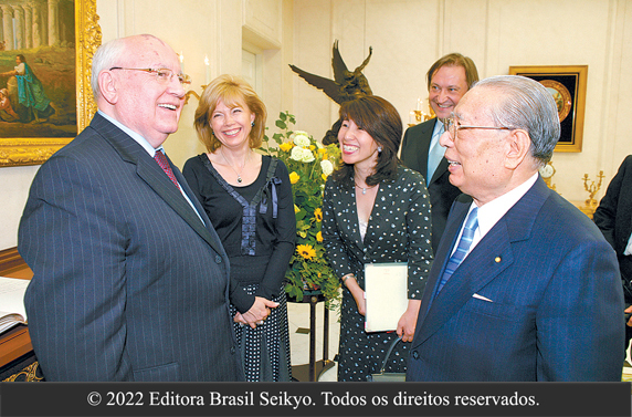 Ao lado de algumas pessoas, presidente Ikeda dialoga alegremente com Mikhail Gorbachev; ambos vestem terno e gravata. No recinto, quadros e estátua integram o ambiente 
