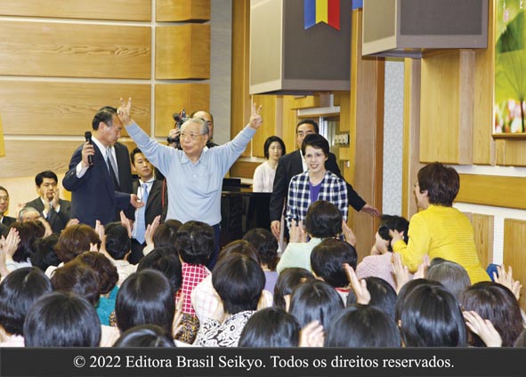 presidente Ikeda de camisa azul clara faz v da vitória com os dedos, erguendo as mãos acima da cabeça, à frente de várias pessoas, dentro de uma sala de reuniões com paredes claras